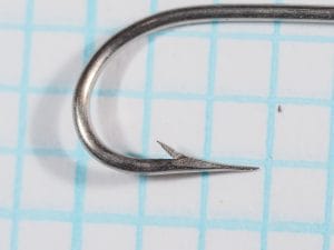 Close view of a hook point and barb on blue grid paper, showing a tapering point with a nearly straight inner face.