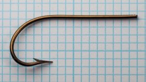 Mustad 3310 size 23 full side profile showing a blind marked shank, rounded bend, barbed point, and bronzed finish on a measurement grid.