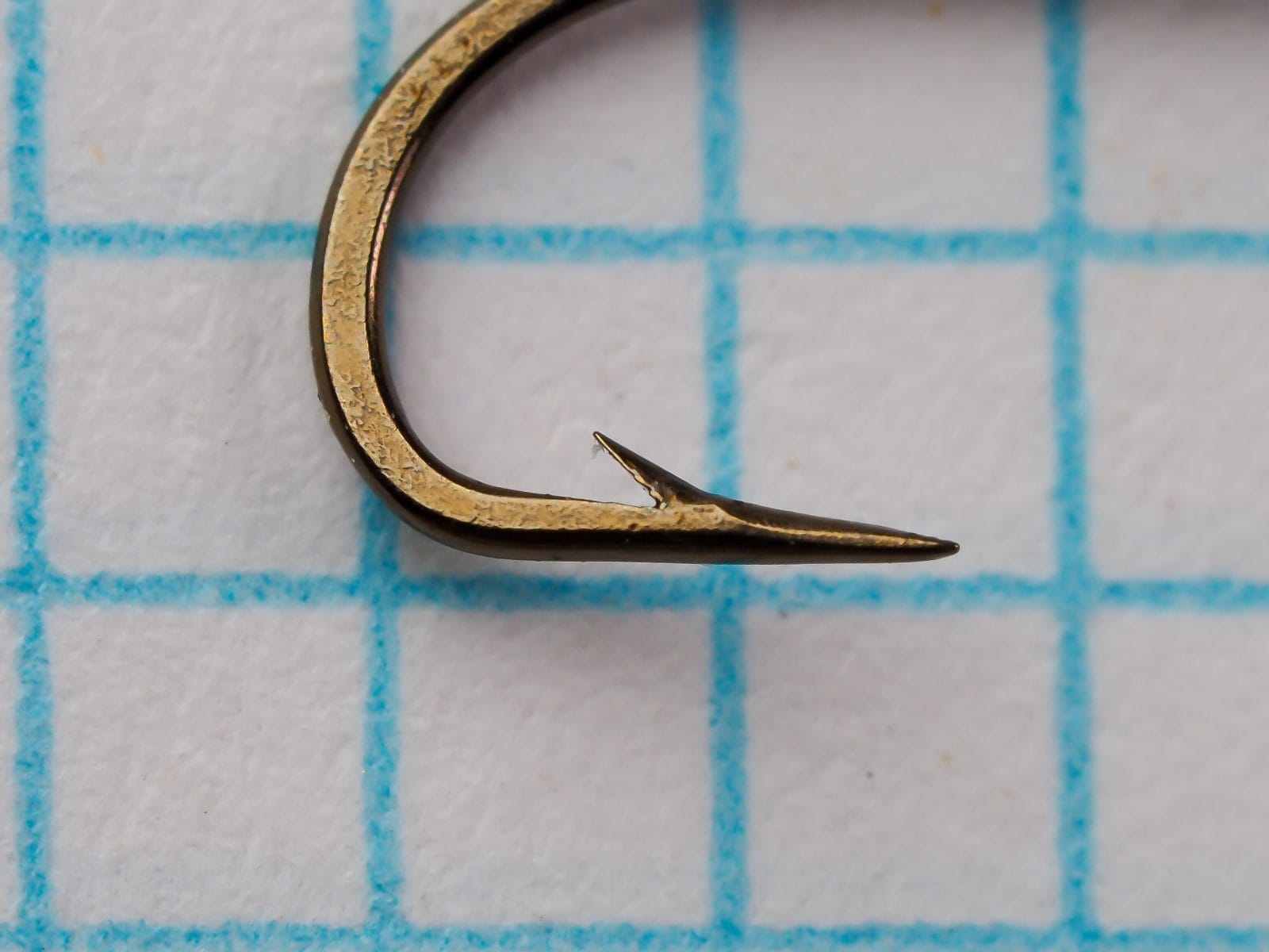 Mustad 56310 — close-up of hook point showing slight reversed offset and barb angle — bronzed finish with minimal patina — on 1/10-inch blue measurement grid