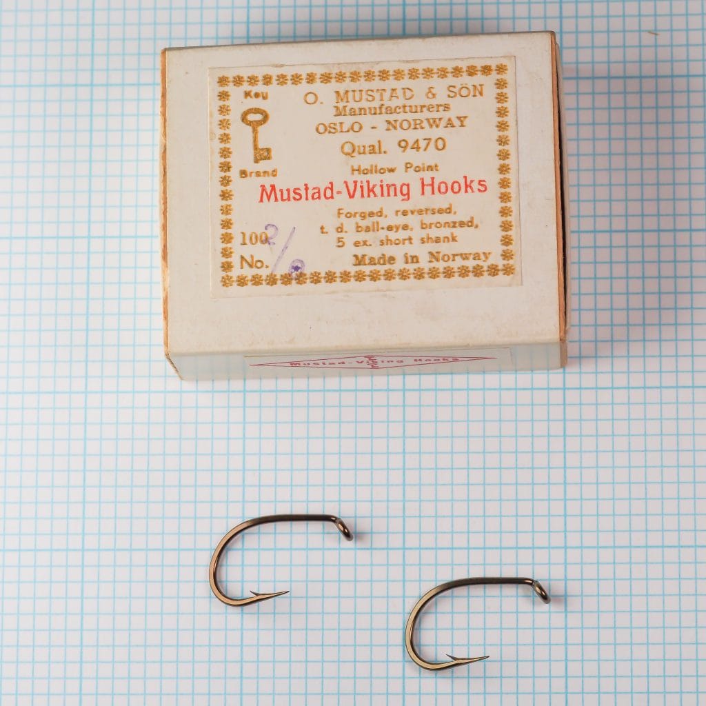 Mustad 9470 box with hooks and label — vintage cardboard slide-box with cream and gold printing, size 2/0, showing closed box and two loose bronzed hooks on measurement grid