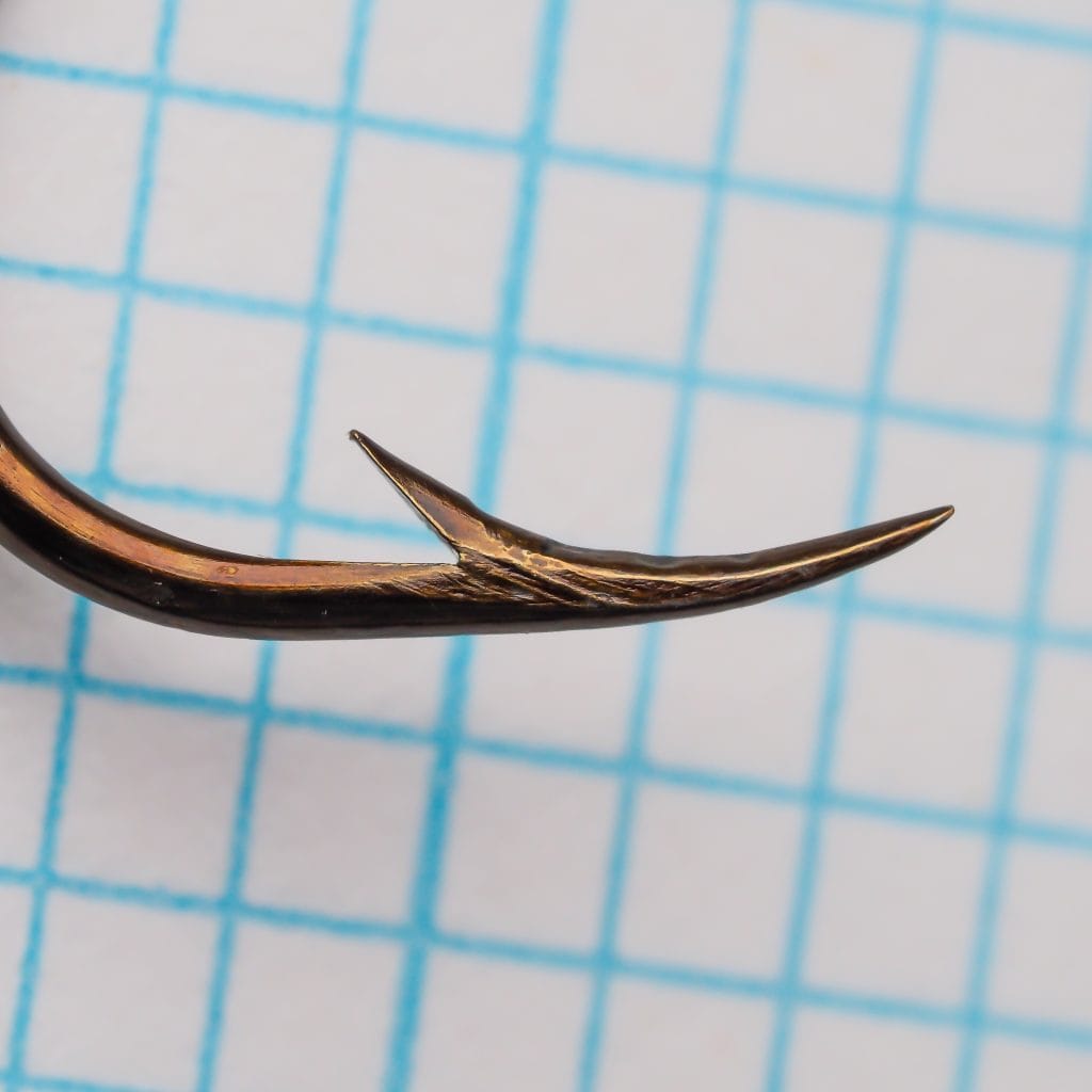 Mustad 9555 Hook Point — close-up detail of the reversed (beak) hollow point showing the inward-curved geometry and sharp tip — size 3/0 — bronzed finish — on measurement grid