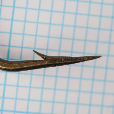 Mustad 9617 — hook point profile — size 6/0 — concave hollow point geometry, sharp tip, bronzed finish — against 1/10-inch measurement grid