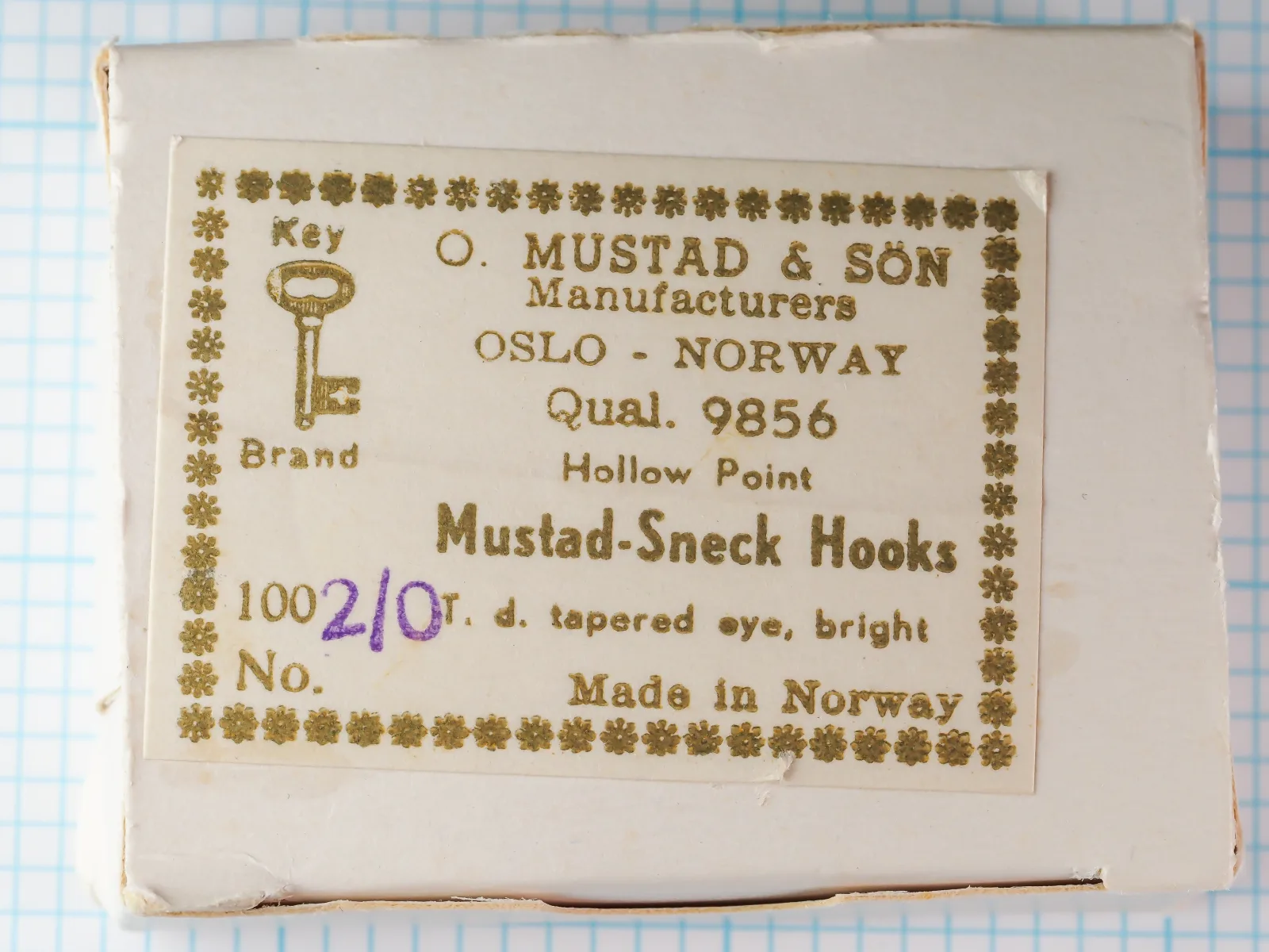 Mustad box lid with a gold-printed Key Brand label reading Qual. 9856, Hollow Point, Mustad-Sneck Hooks, with size 2/0 written in purple.