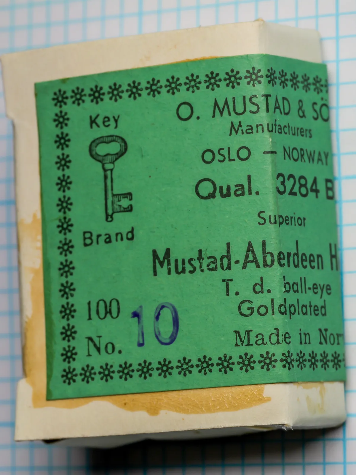Upright green Mustad label with a key drawing, printed lines and a blue handwritten 10.