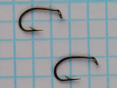 Mustad 543 H size 17 — two specimen hooks photographed from above — showing rounded sproat bend, short shank, tapered turned-down eye, and offset (kirbed) point — bronzed finish — on 1/10-inch measurement grid