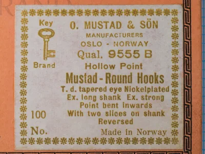 Mustad 9555 B label close-up — gold letterpress text on cream background — full eight-line specification including Hollow Point, Round Hooks, tapered eye, nickelplated, extra long shank, extra strong, point bent inwards, two slices, reversed