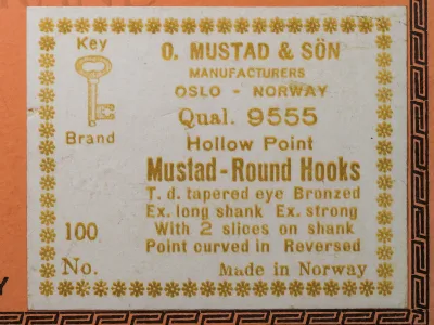 Mustad 9555 Sales Card Back Label — gold-ink printed specifications detailing tapered eye, bronzed finish, extra-long shank, extra-strong wire, bait-gripping slices, curved-in reversed point, and 'Made in Norway' designation