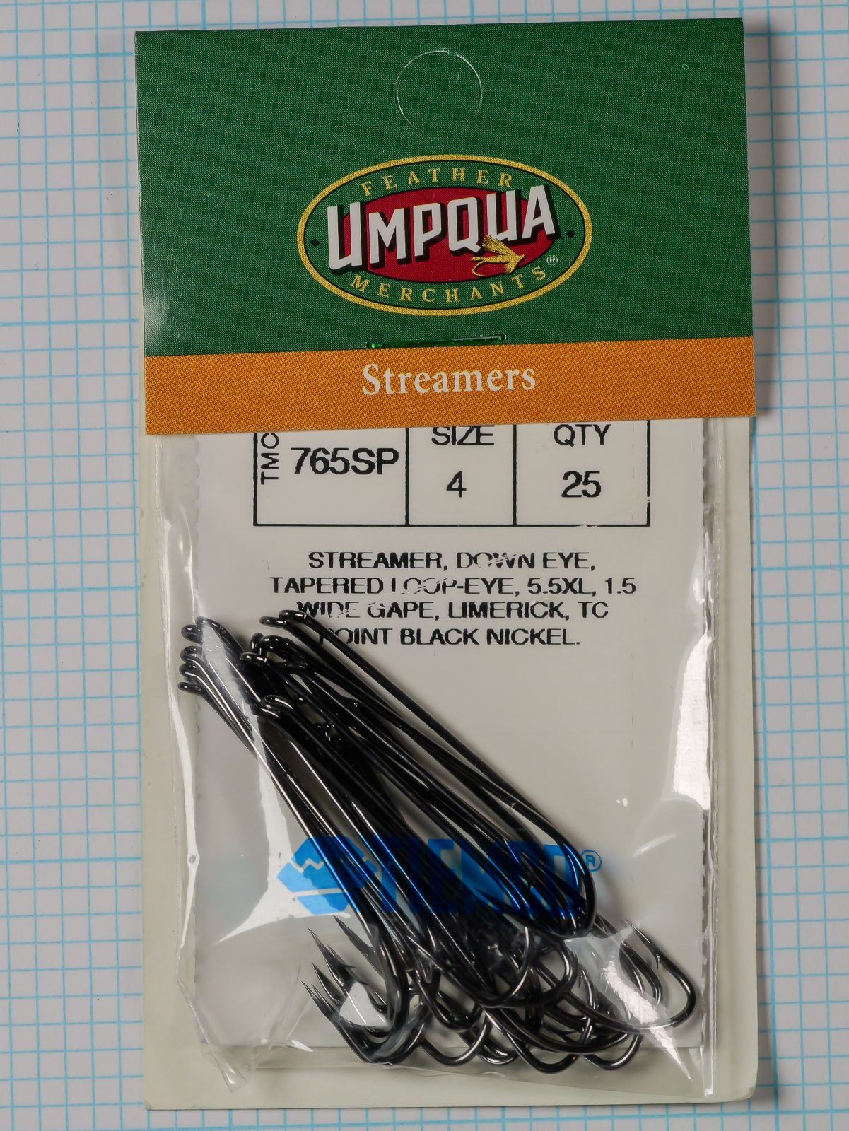 Tiemco TMC 765SP Streamer — clamshell package front showing Feather Umpqua Merchants branding and Size 4 qty 25 specification label with 25 black nickel hooks visible inside sealed packaging on measurement grid