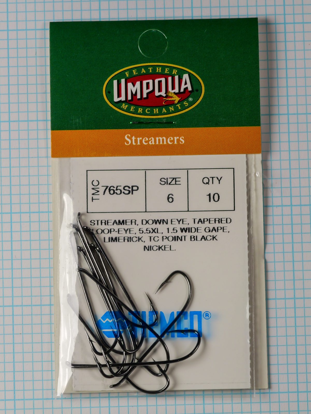 Tiemco TMC 765SP Streamer — clamshell package front showing Size 6 qty 10 specification label with black nickel hooks visible inside opened packaging on measurement grid