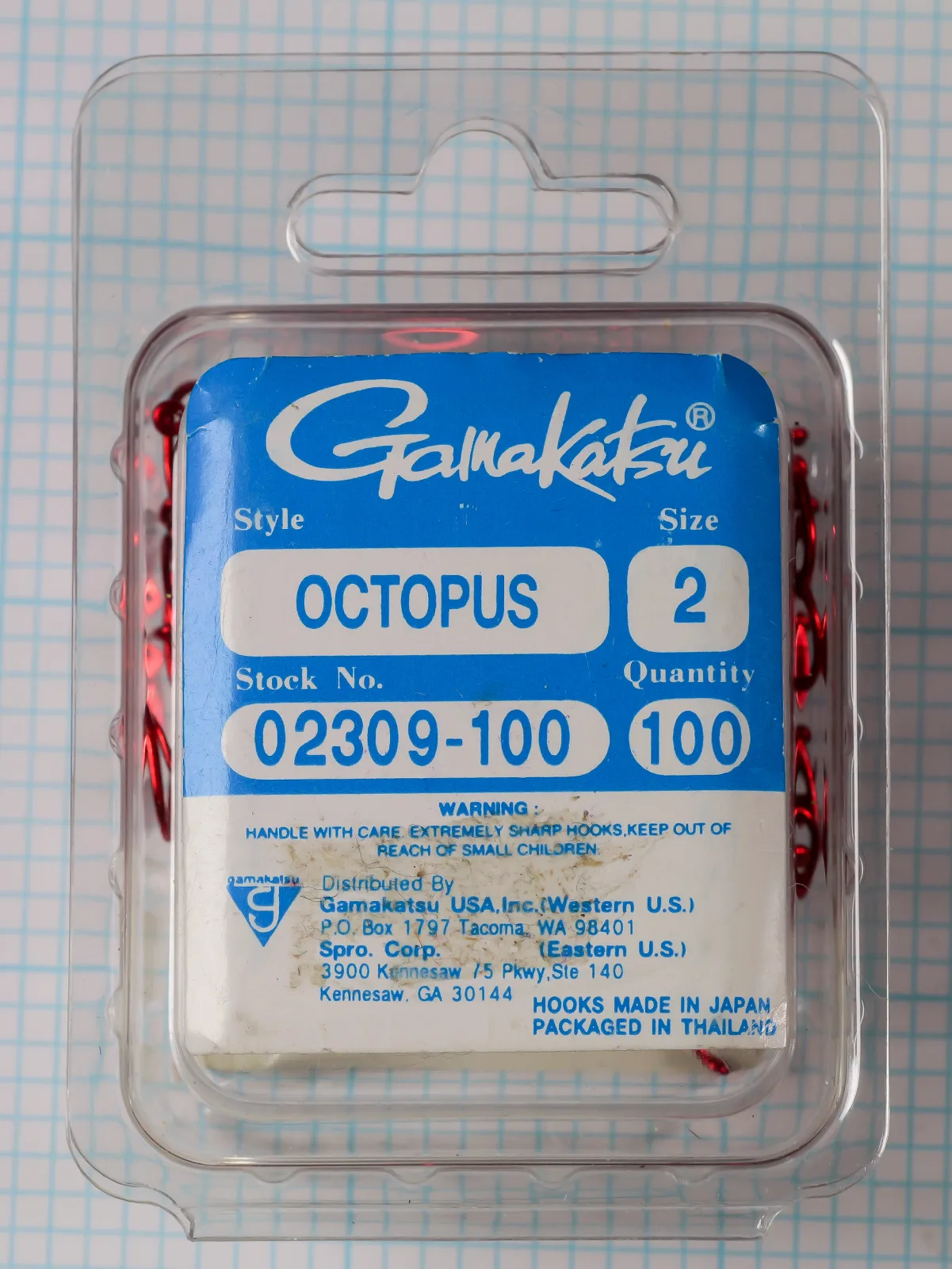 Gamakatsu 02309 Octopus Hook Size 2 — packaged blister card with blue label on measurement grid — shows complete clamshell packaging with die-cut hanging hole