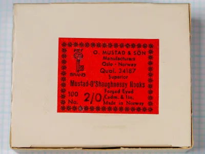 Mustad 34187 O'Shaughnessy box exterior — cream-colored board with red label showing anchor-and-crown logo and letterpress text — 100 hooks, size 2/0, tinned finish