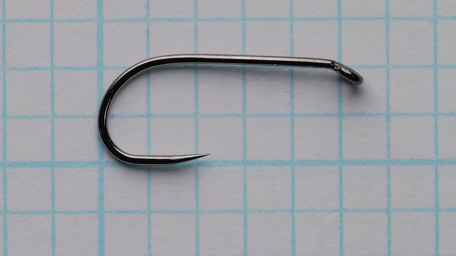 Side view of a dark Ahrex FW503 size 12 hook on blue grid paper, with a closed ring eye at the right, a plain shank, a rounded bend at the left and a barbless point.