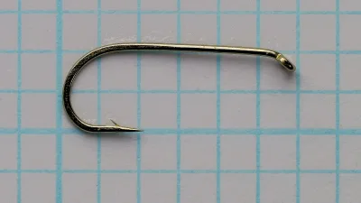 Gold-bronze fish hook lying on a blue grid, eye at right turned down, round bend at left, barbed point aimed back toward the eye.