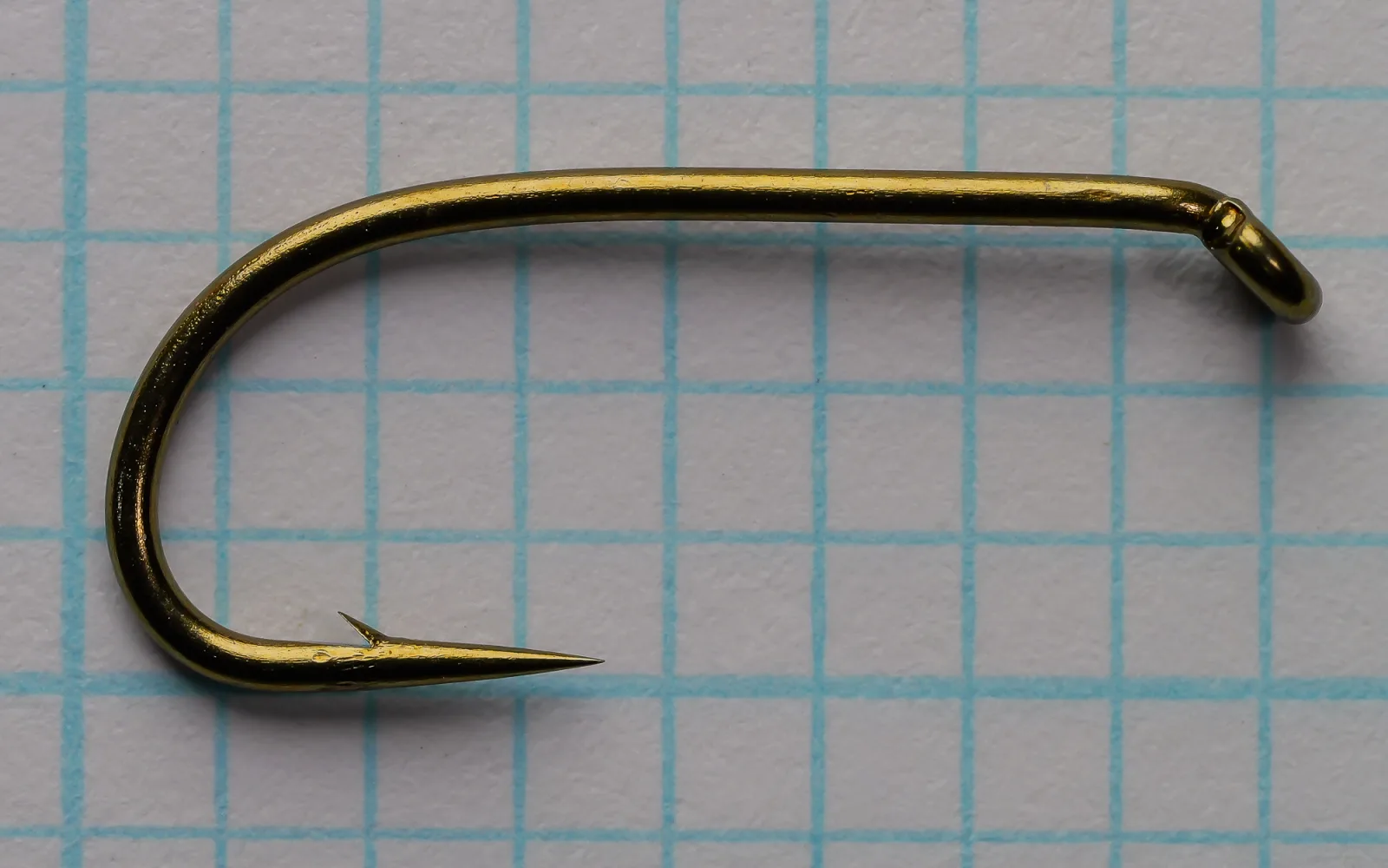 Bronze fly hook with a straight shank, down-turned eye on the right, rounded bend, and barbed point on the left, photographed flat on blue grid paper.