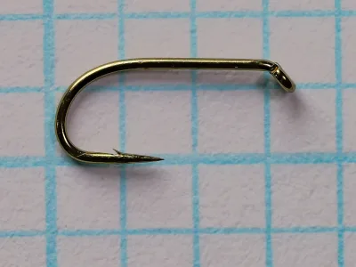 Side view of a bronzed fly hook with a round bend, straight plain shank, closed turned-down eye, long tapered point and small barb, lying on blue grid paper