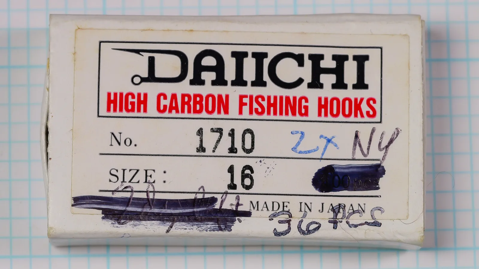 White paper-wrapped box with a pasted lid label reading DAIICHI, HIGH CARBON FISHING HOOKS, No. 1710, SIZE : 16 and MADE IN JAPAN, with pen marks over the quantity, on blue grid paper.