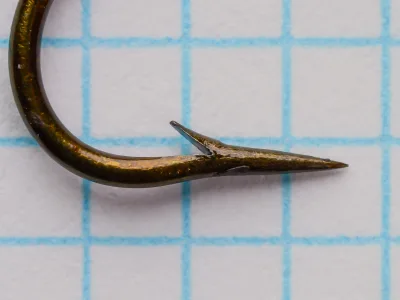 Close-up of a hook point on a grid background, showing a long tapering point with file facets and a barb cut on the gap side.