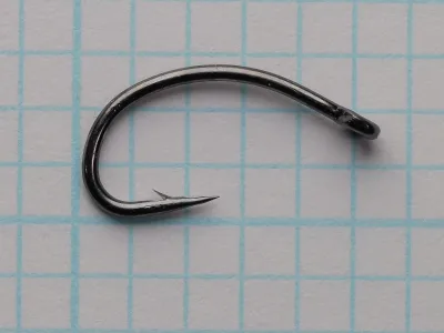 Dark glossy fly hook with a curved shank, deep round bend, barbed point and small closed eye, lying on blue grid paper in side view.