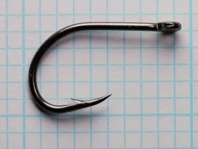 Side view of a black fishhook with a straight plain shank, a deep round bend, a wide gap and one cut barb, lying flat on blue graph paper.