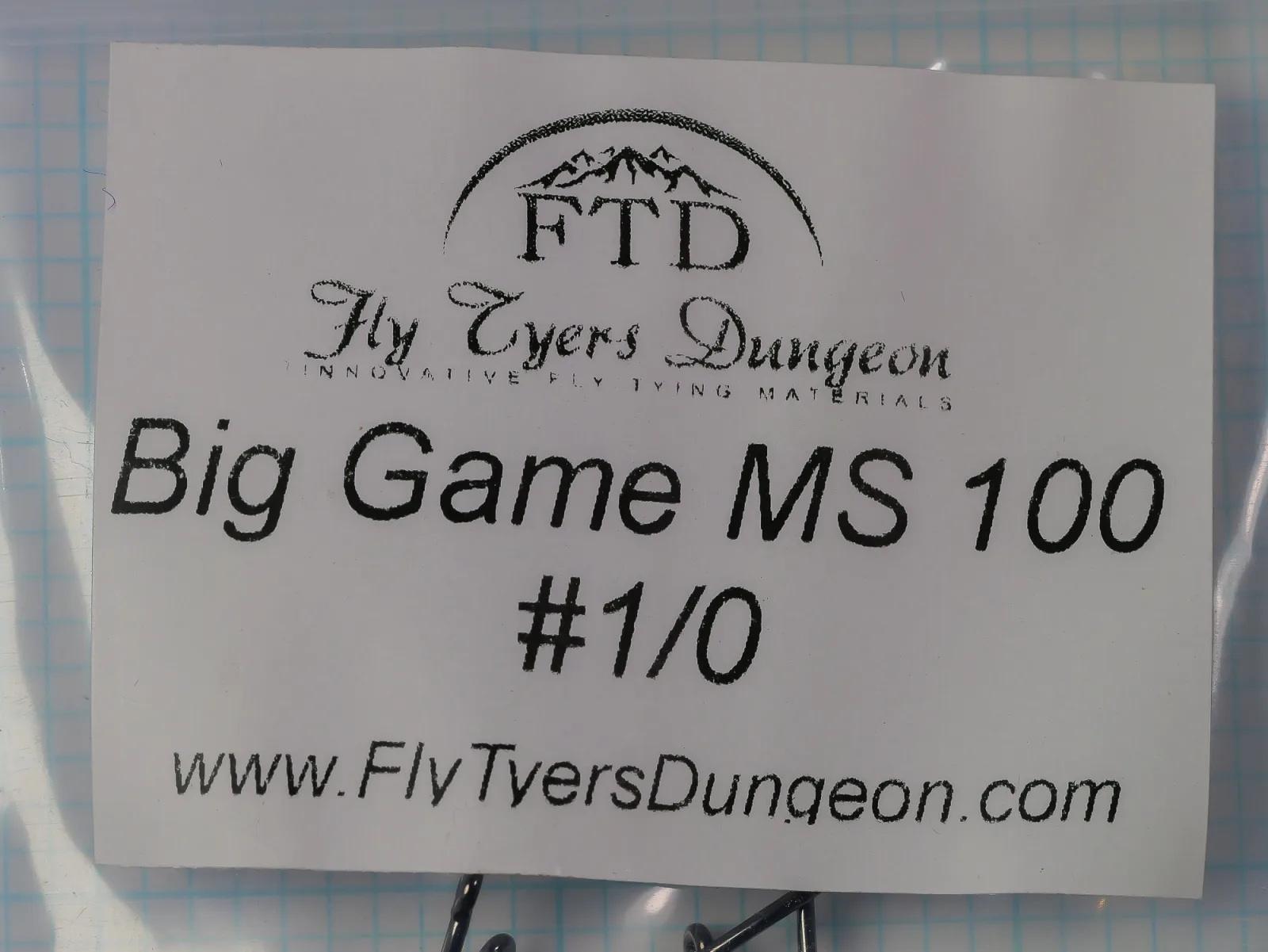 White paper label on a clear plastic bag reading Big Game MS 100 and #1/0, with an arched FTD logo and a Fly Tyers Dungeon wordmark.