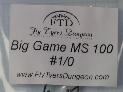 White paper label on a clear plastic bag reading Big Game MS 100 and #1/0, with an arched FTD logo and a Fly Tyers Dungeon wordmark.