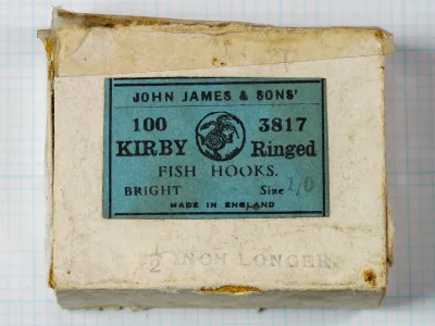 Lid of a small cardboard box with a blue label reading John James & Sons' Kirby Ringed fish hooks, 100, 3817, and a stamp below it reading 1/2 inch longer.