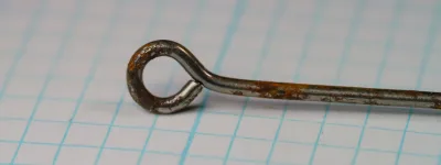 Close view of the ring eye of a rusted hook on grid paper, the wire bent into a loop with a narrow seam at the wire end.