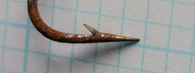 Close view of a rusted hook point with a single cut barb on its inner side, on grid paper.