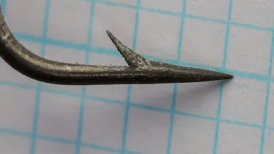 Close view of a hook point on graph paper, tapering to a fine tip with a single short barb on the shank side.