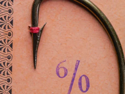 Close-up of the 6/0 hook's long straight point and slender barb, sewn to the card with red thread beside its purple 6/0 stamp.