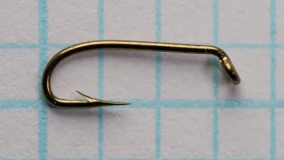 Side view of a bronzed Mustad 3906B size 18 hook with a turned-down eye, on blue grid paper.