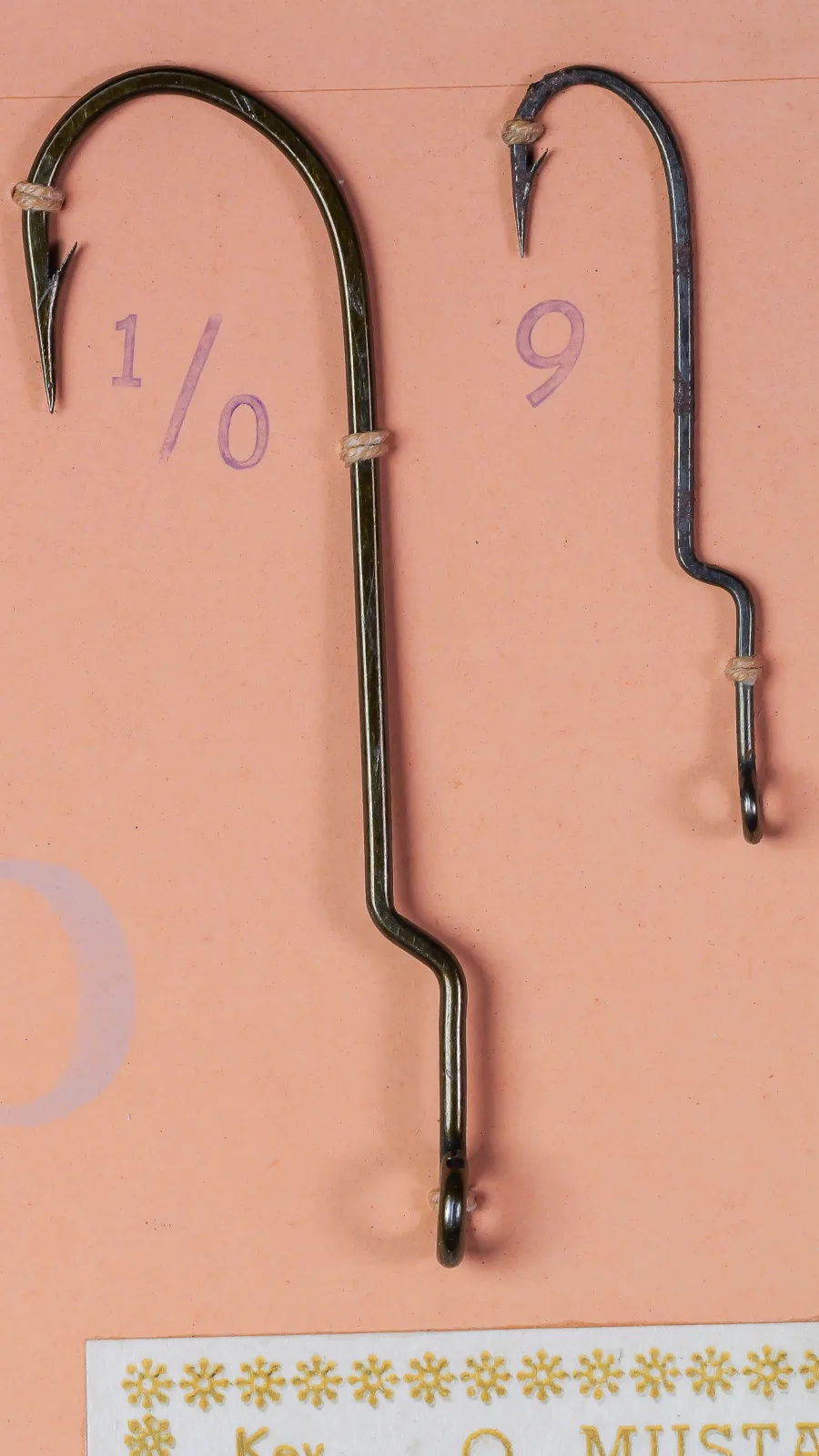 Close view of two Mustad 7959 A hooks tied to an orange card, stamped 1/0 and 9, each with a straight ring eye and a crank near the eye end of the shank.