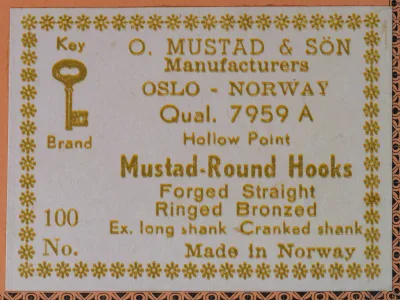 Gold-printed white label with a key emblem, reading O. Mustad & Sön, Qual. 7959 A, Hollow Point, Mustad-Round Hooks, Forged Straight, Ringed Bronzed, Ex. long shank Cranked shank, Made in Norway.