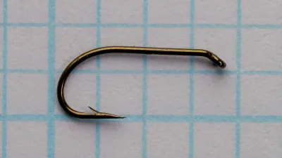 Side view of a size 16 hook with a turned-down eye, plain straight shank, round bend, barbed point, and dark bronze-gold finish, lying on grid paper.