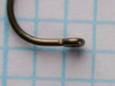 Close view of the point end of a size 10 Tag hook showing a flattened, rounded head with a hole and a small nick behind it, on grid paper.
