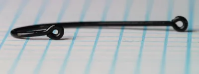 Size 12 hook photographed at a low angle on grid paper, black coating, with a closed eye at the shank end and a second closed eye at the end of the bend arm.