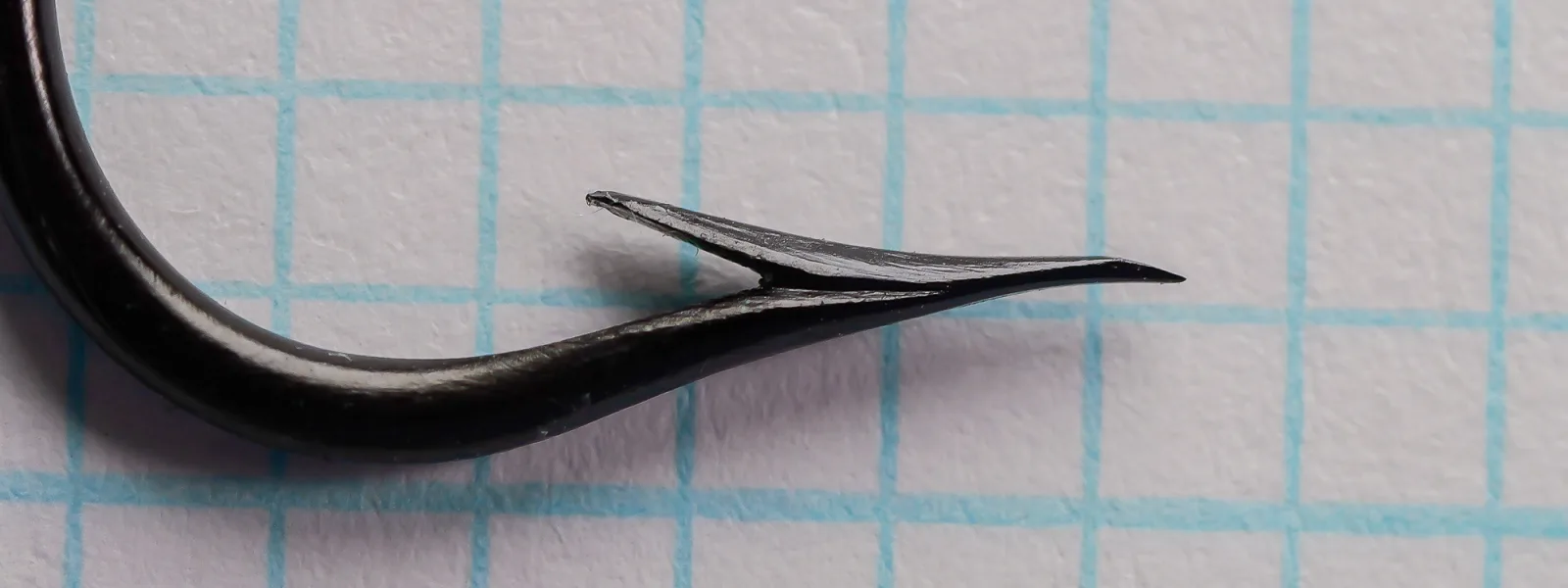 Close view of the point and barb of a black blind-eye hook on blue grid paper, showing a large raised barb set off by a narrow V notch.