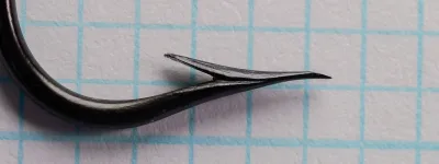 Close view of the point and barb of a black blind-eye hook on blue grid paper, showing a large raised barb set off by a narrow V notch.